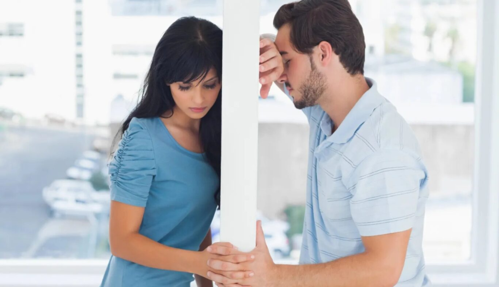 A man and a woman stand separated by a partition, appearing upset or deep in thought.