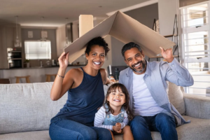 A happy family sitting on a couch, holding a cardboard roof over their heads, symbolizing parenting as a team.